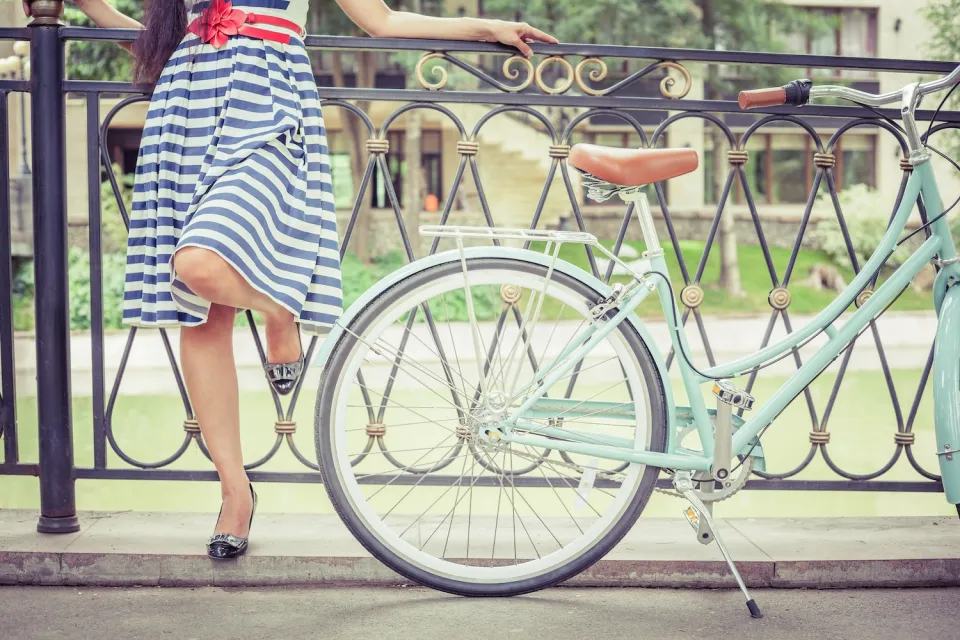 Close-up on woman in white and blue striped knee-length dress standing next to mint green cruiser bike resting against low wrought iron fence in front of green lawn.